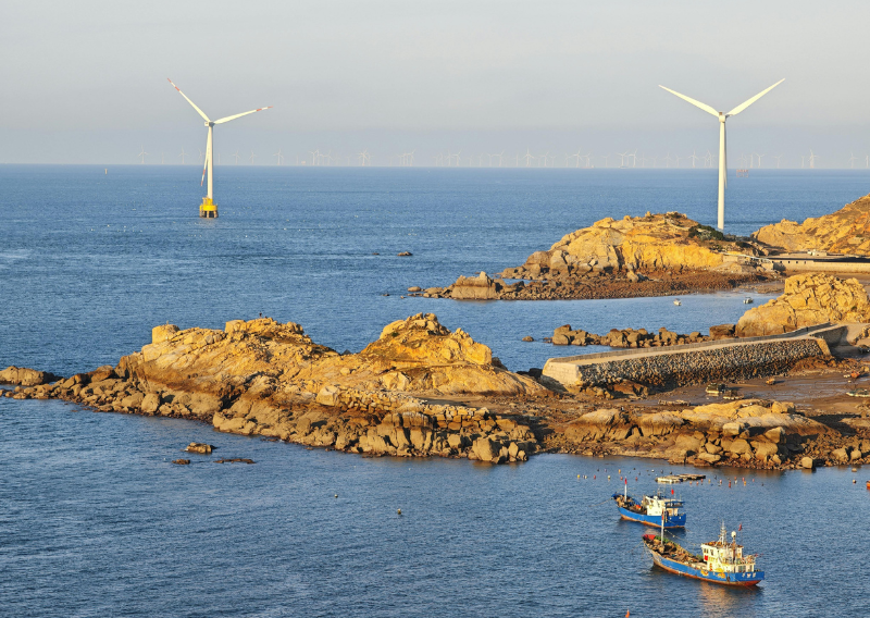 Offshore wind turbines along a rocky coastline with fishing boats at sea, symbolising global energy markets themes relevant to forex trading in Singapore.